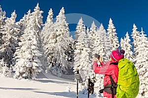 Caucasian young woman in colorful outfit taking a break from backcountry skiing taking a picture, recording the beautiful winter