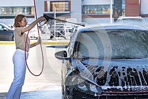Caucasian young woman driver washing at manual car washing, cleaning with foam, pressured water. Self-service car wash with high-p