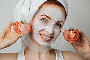 Caucasian young woman holding slices of tomato while posing in studio with white clay mask