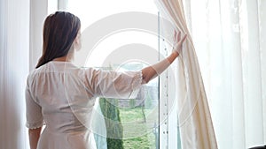 Caucasian young woman opening curtain and looking through window.