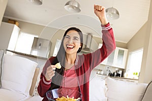 Caucasian young woman raising hand and screaming cheerfully while having chips on sofa at home