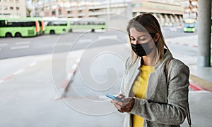 Caucasian young woman with smartphone waiting at bus station while wearing safety mask for coronavirus outbreak - Focus on girl`s