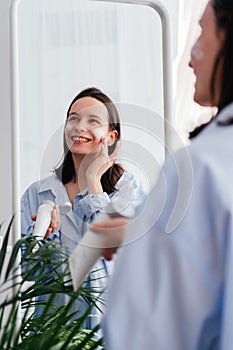 Caucasian young woman touching face and applying cream looking in mirror indoors. Home beauty skincare concept