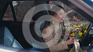 Caucasian young woman walking to car sitting down on driver's seat smelling bouquet. Smiling happy confident lady