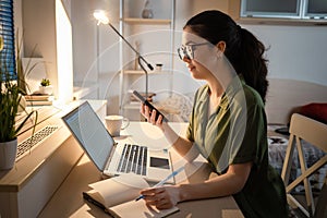 Caucasian young woman wearing glasses using cellphone and writes in notebook. Side view. Concept of remote work and