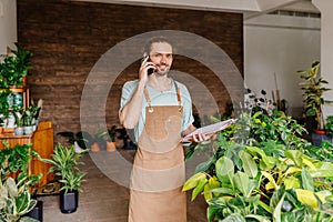 Caucasian young worker man in apron holding potted flower and talking on mobile phone during work in greenhouse