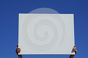 Causasian woman is holding a blank white canvas in her hands on a sunny day with blue cloudless sky in background.