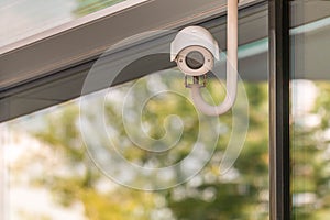 CCTV or surveillance camera operating on window building with green trees foliage reflection of shopping mall