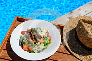 Ceasar salad with shrimps served on wooden tray with straw hat on left and tropical water in background