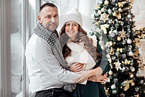 Celebrating Christmas together. Young couple in holiday clothes hugging near the Christmas tree. New Year, Valentine, love,