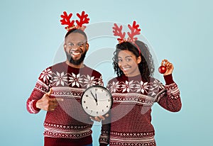 Celebrating New Year. Happy black spouses with clock showing five minutes till midnight, wearing deer horns and sweaters