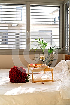 Celebrating Saint Valentine`s Day with bouquet of red roses, glass of champagne and croissants on tray on bed. Romantic surprise