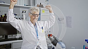 Celebrating a win, a senior grey-haired woman scientist is all smiles while working at her lab microscope, brimming with