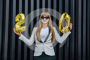 Celebration concept with golden balloons. Young woman in suit holding air balloons on grey background