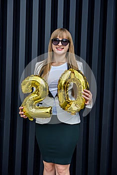 Celebration concept with golden balloons. Young woman in suit holding air balloons on grey background