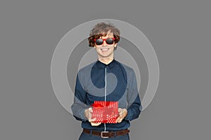 Celebration Concept. Teenager boy in sunglasses standing isolated on grey with present smiling cheerful