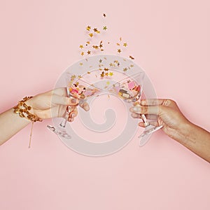 Celebration and holiday background. Two hands holding wine glasses with gold confetti on pink background