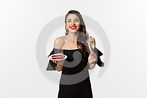 Celebration and party concept. Happy woman holding birthday cake and drinking champagne, smiling while standing in black