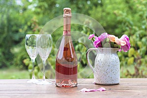 Celebration still life. Bottle of pink champagne, two glasses and bouquet of pink roses in rutsitc jug on wooden table in green