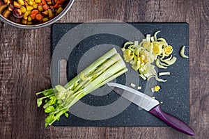 Celery stalks, with some chopped, on a black cutting board, chef knife, wood table, bowl of diced rainbow carrots