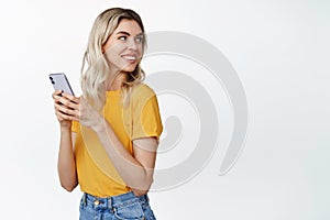 Celluar technology. Young blond woman holds mobile phone, turn head behind and smile at copy space, stands in yellow