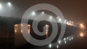 Cement bridge in thick fog. Street lights illuminate road at night, reflecting in calm dark river water. Mystical atmosphere.