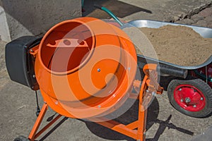 Cement mixer machine is at construction site with wheelbarrow, tools, sand and cement bag. Orange concrete mixer at building