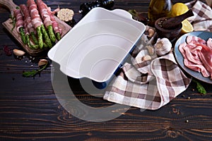 ceramic baking dish for cooking aspargus covered with bacon on wooden kitchen table