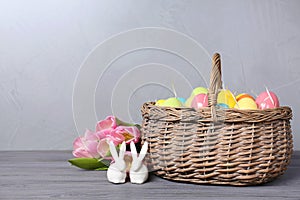 Ceramic bunnies near wicker basket with Easter eggs and spring tulips on wooden table against light background