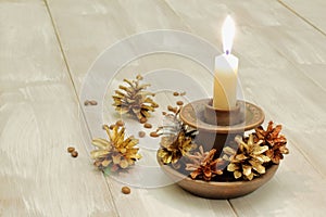 Ceramic candlestick and burning white wax candle, multicolored pine cones and coffee grains on light wooden background