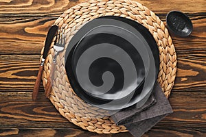 Ceramic empty black plate, silverware and linen kitchen towel napkin on old brawn wooden table background. Cooking stone backdrop