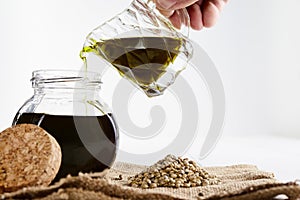 Ceramic jar pouring virgin vegetable oil in a glass bottle and hemp seeds on sackcloth. close up