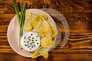 Ceramic plate with potato chips and glass bowl with sour cream on wooden table. Top view