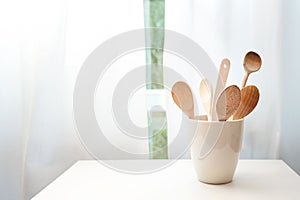 Ceramic pot with wooden cooking spoons on a table in front of the kitchen window with white curtains, copy space