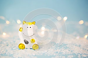 Ceramic toy white cow with gold spots-a symbol of the new year 2021. Christmas blue background with garland lights in blur, copy