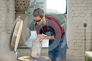 Ceramist Dressed in an Apron Placing Clay Sculpture in Electric Oven. photo