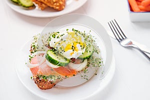 Cereal bun with egg benedict, smoked salmon, sprout micro greens, onion and cucumber slices, cream cheese on the served white wood