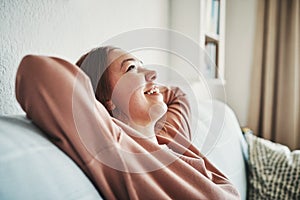 Cest la vie. an attractive young woman sitting alone in her living room with her hands behind her head.