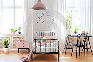 Chair at desk next to bed in pink and white girl`s room interior with plants and windows