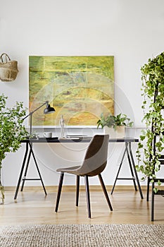 Chair at table with lamp in modern home office interior with plants and painting. Real photo