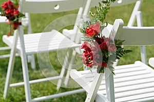 Chairs decorated with fresh flowers in red tones for the wedding ceremony on the green lawn