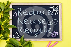 Chalk board Reduse Reuse Recycle sign on a yellow background with green leaves and chalk pieces. Top view