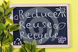 Chalk board Reduse Reuse Recycle sign on a yellow background with green leaves. Top view