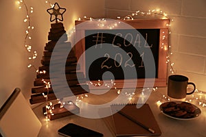 Chalkboard with hashtag Goal 2021, fairy lights near notebooks, smartphone and cookies on white table