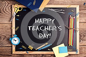 A chalkboard with the text happy teachers day written in it, school supplies and alarm clock on a rustic wooden table