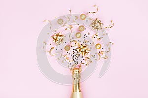 Champagne bottle with christmas decoration from confetti stars, golden balls and party streamers on pink background. Flat lay.