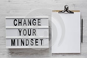 `Change your mindset` words on a lightbox, clipboard with blank sheet of paper on a white wooden surface, overhead view. Top vie