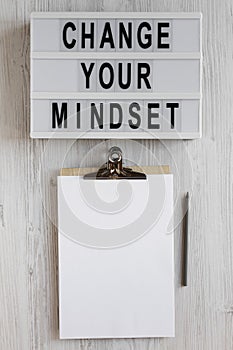 `Change your mindset` words on a modern board, clipboard with blank sheet of paper on a white wooden background, overhead view.