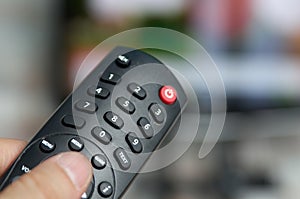 Changing TV program: close up of human hand holding television remote control on blured background with TV screen