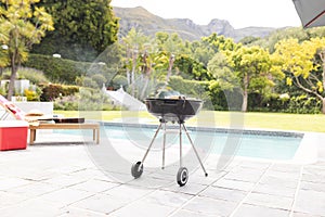 A charcoal grill is ready for a barbecue by a poolside with lush greenery in the background